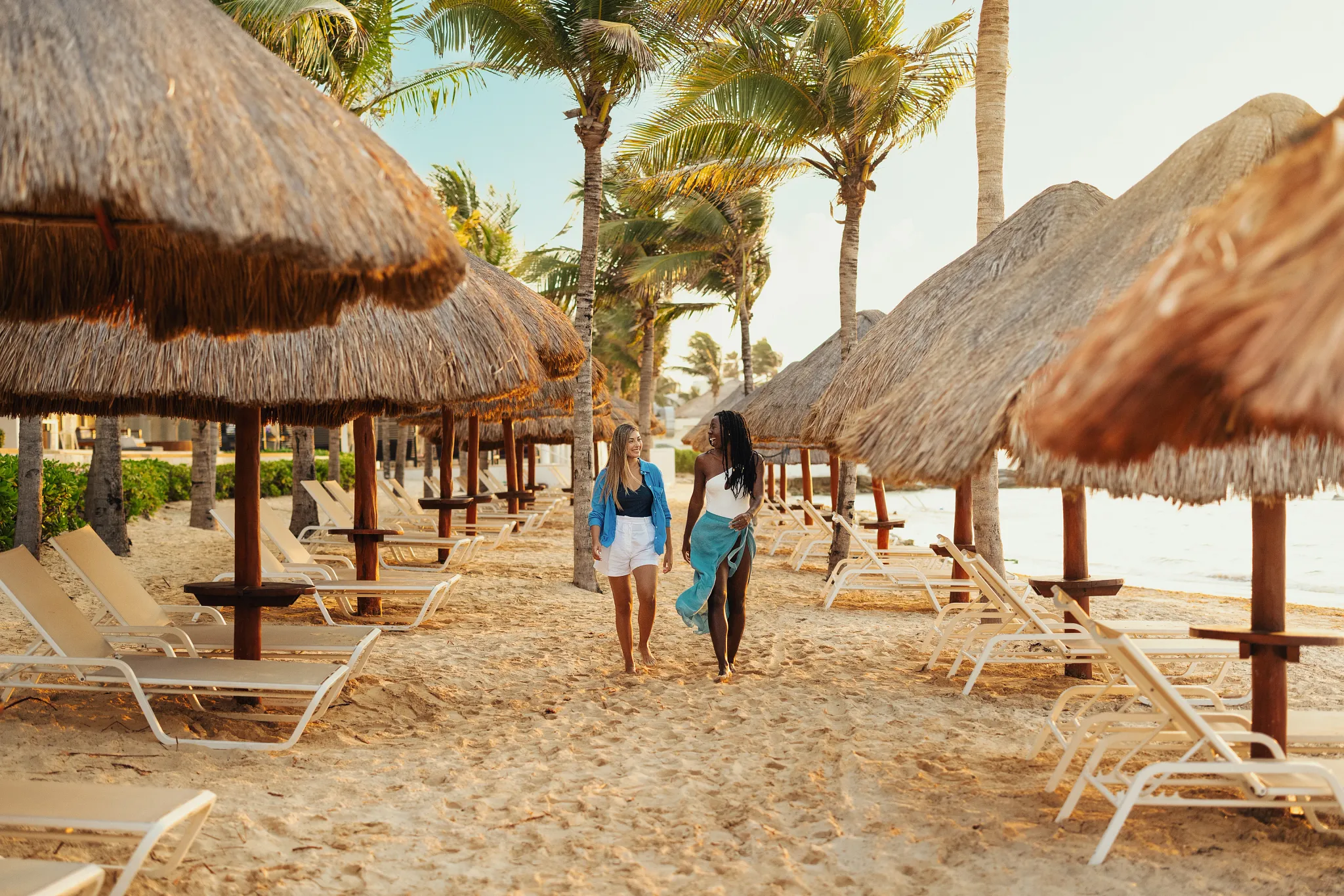 Two women walking under palapas along the sandy beachfront at Breathless Riviera Cancun, an adults-only all-inclusive resort in Mexico.
