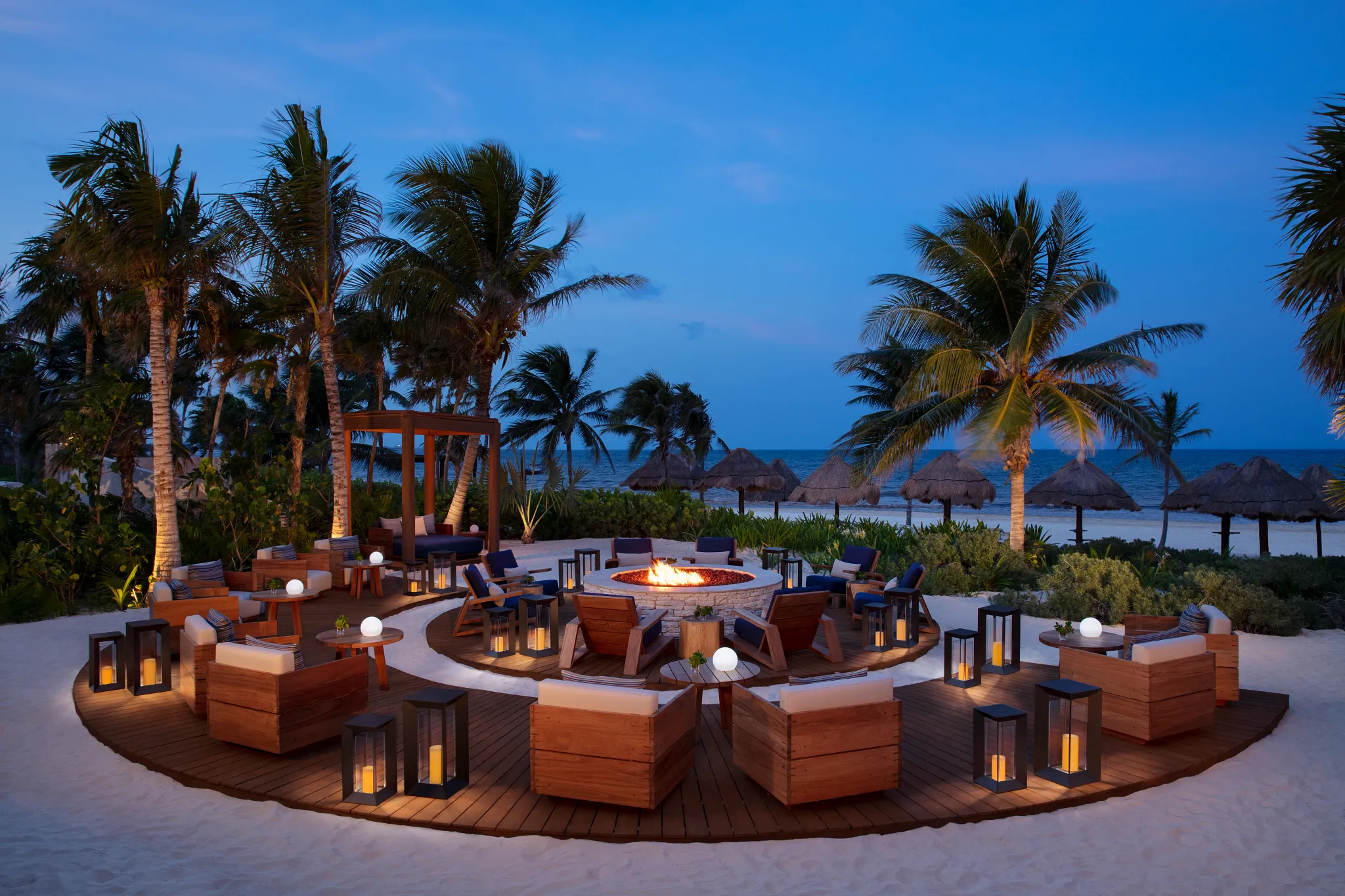 Beachfront fire pit lounge at Secrets Maroma Beach Riviera Cancun surrounded by palm trees and ocean views at dusk.
